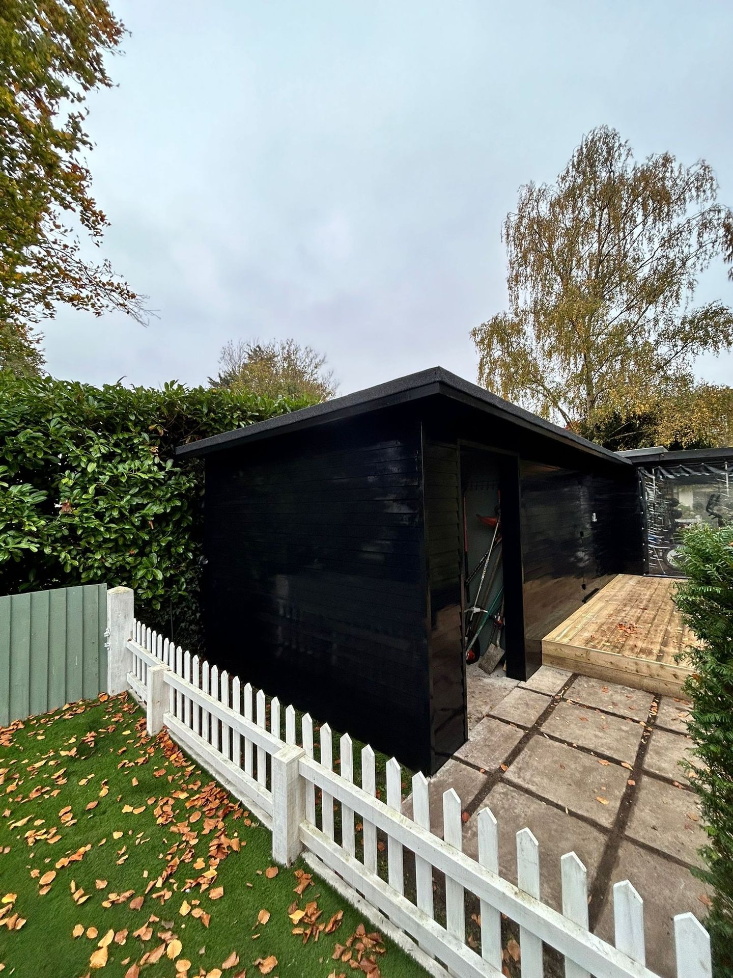 Black garden shed with a white picket fence and autumn leaves on the grass.